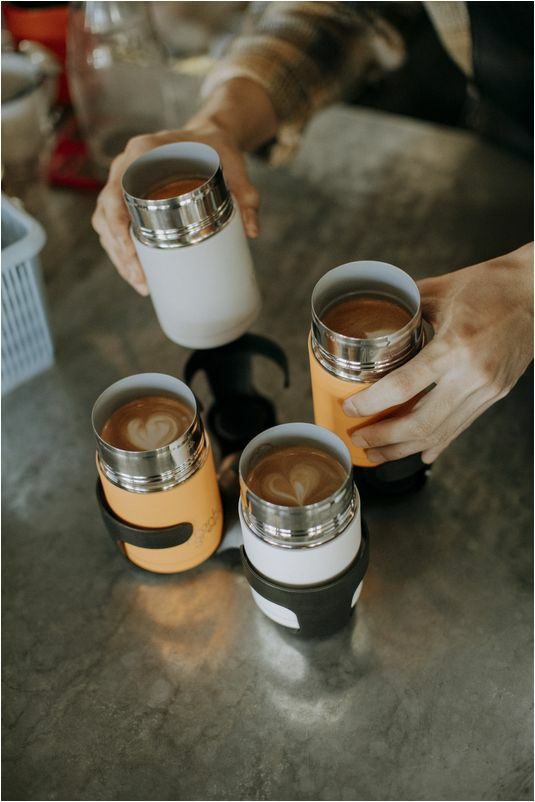 ajándék ötletek férfiaknak: barista szállítható fekete pohártartóba helyez négy nyitott termosz bögrét kávéval, szín alakú hab mintával